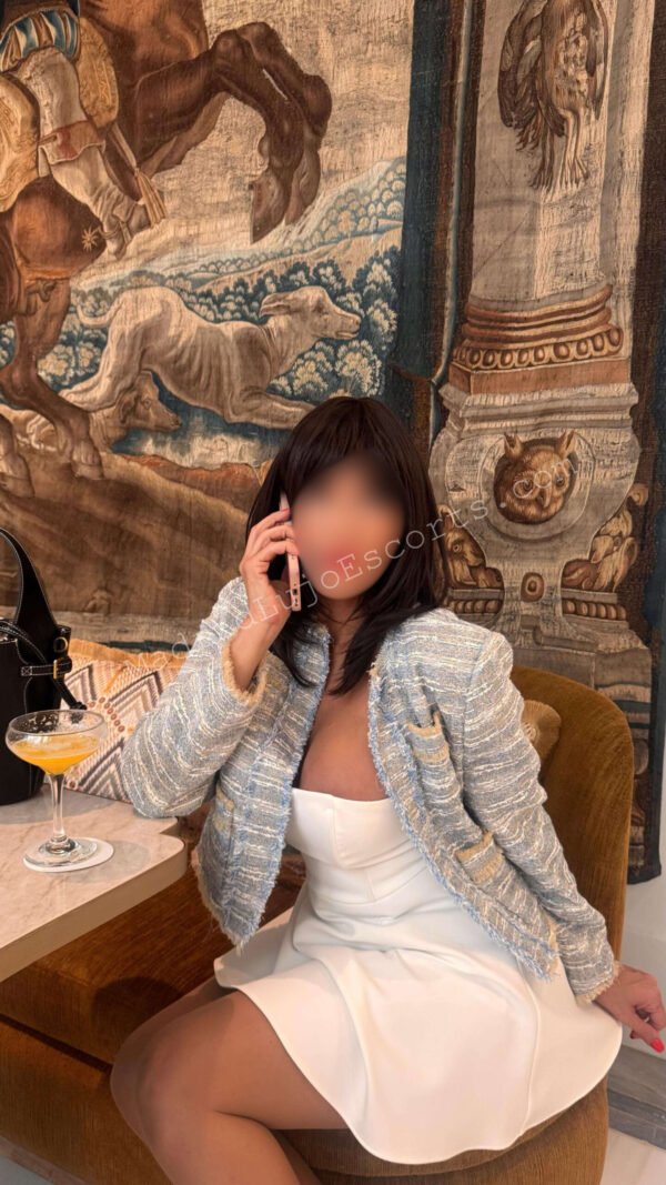 Vanessa en un rincón con vestido blanco y abrigo hablando por teléfono