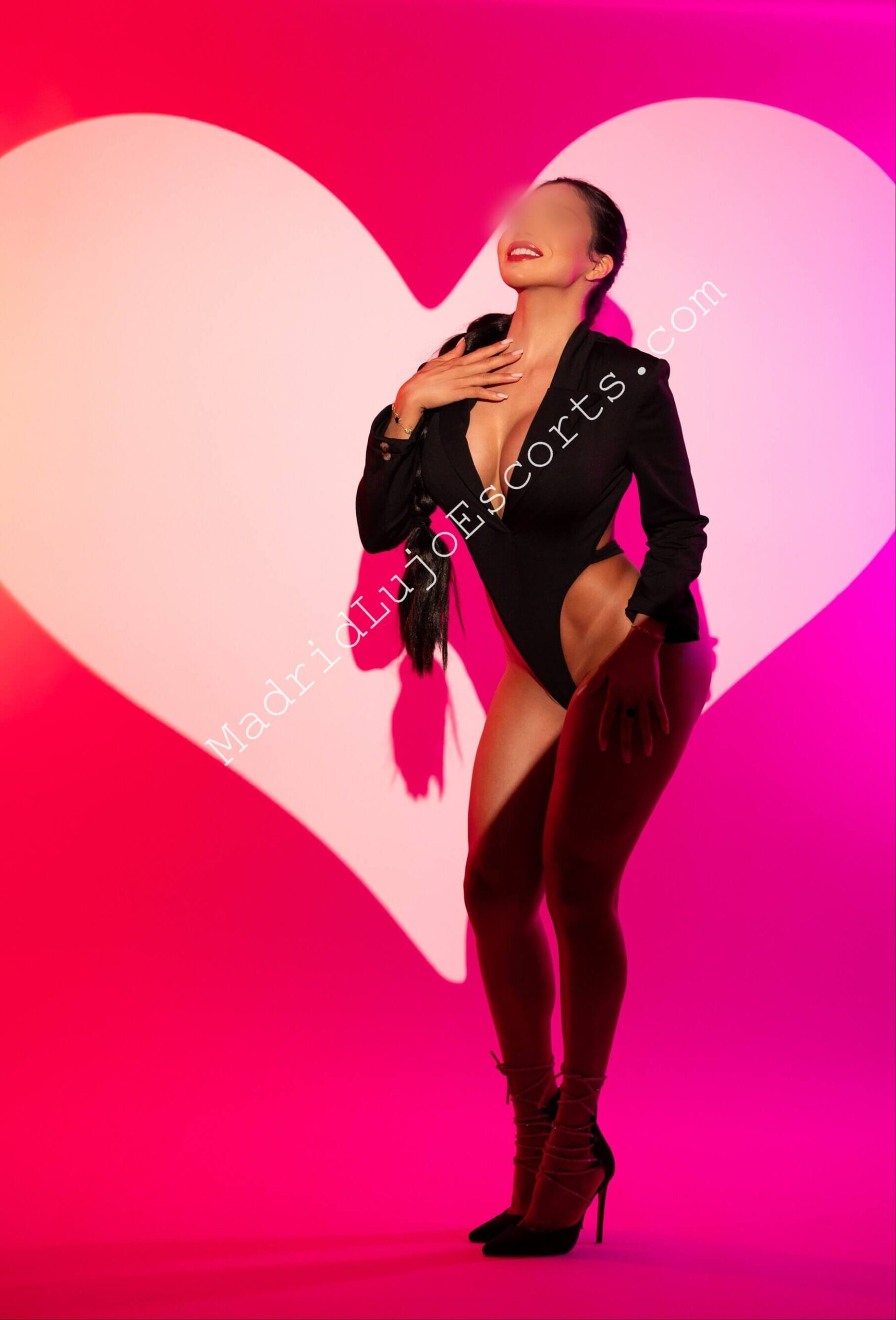 Danielle escort brasileña en Madrid posando con blazer negro frente a fondo rosa con corazones