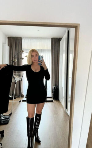 Sara escort española de lujo en Madrid con vestido negro corto y botas altas frente al espejo